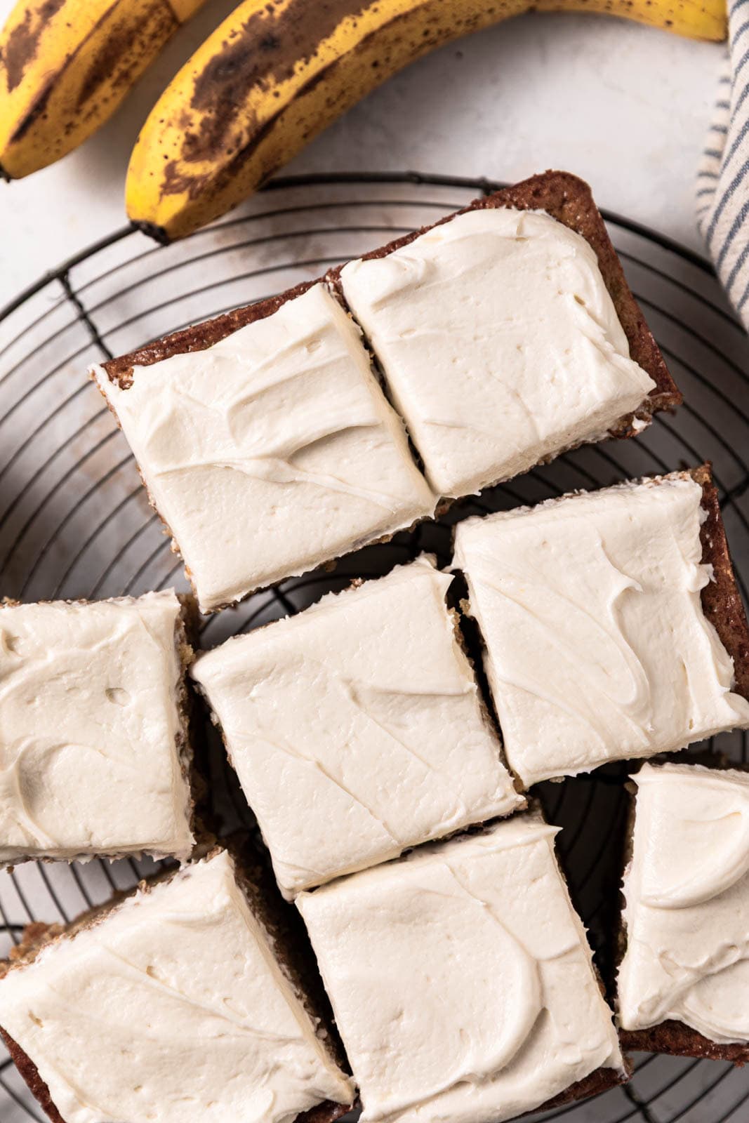 Brown sugar cream cheese frosting spread over banana cake cut into squares on a round wire rack.