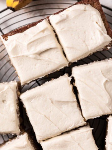 Brown sugar cream cheese frosting spread over banana cake cut into squares on a round wire rack.