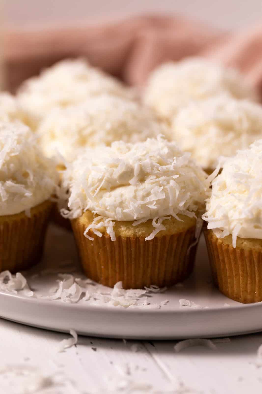 Coconut cupcakes sprinkled with coconut flakes on a round plate set on a white wood board.
