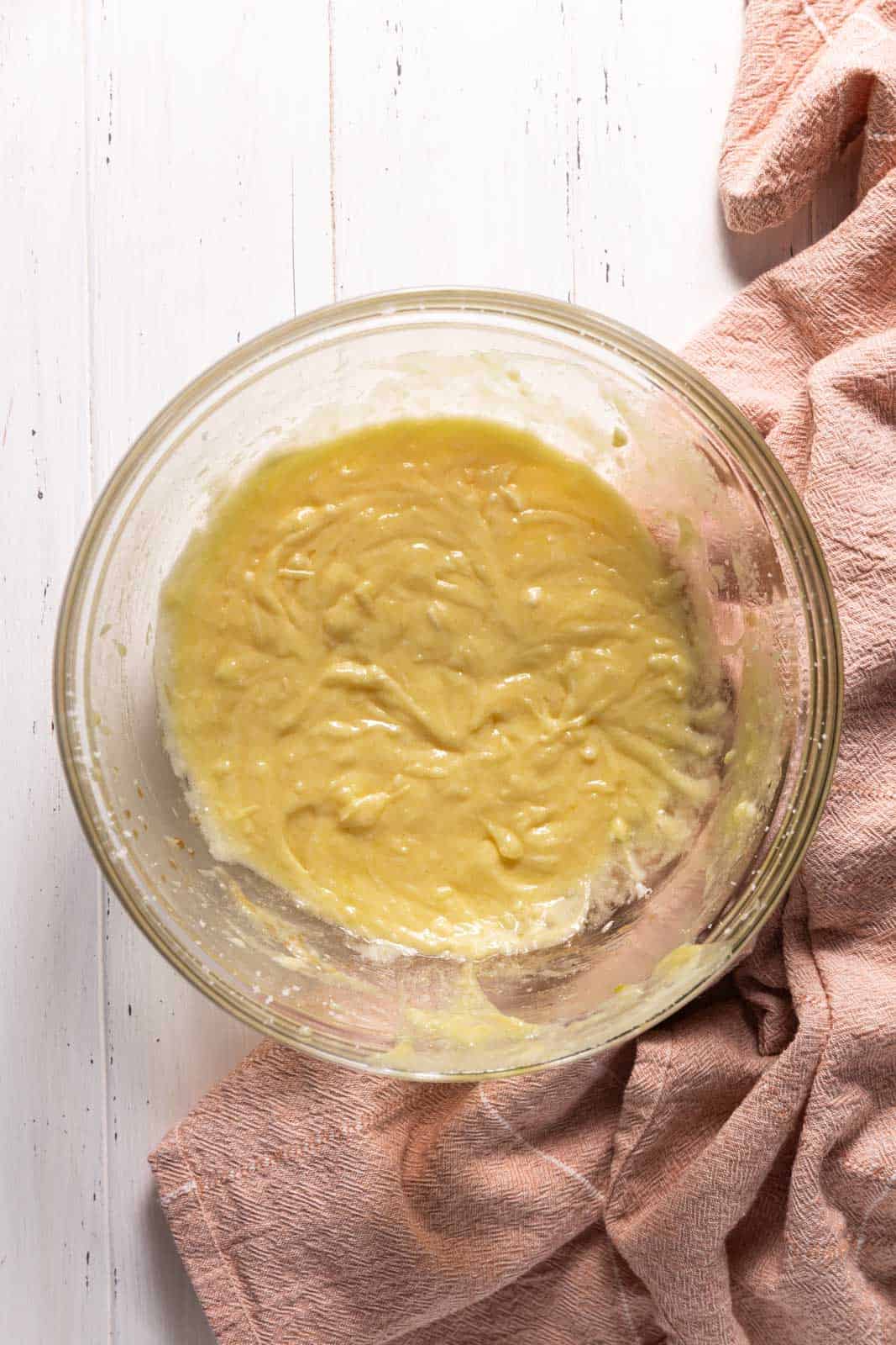Sour cream mixed into creamed butter and sugar mixture with eggs and vanilla in a glass mixing bowl set on a flat surface.