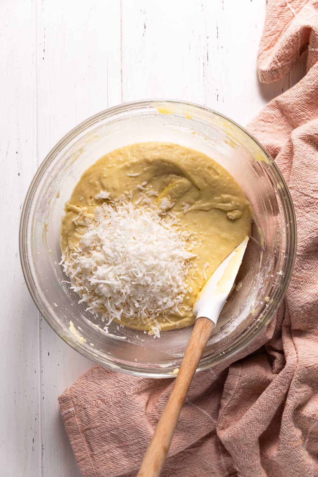 Shredded coconut with coconut cupcake batter in a glass mixing bowl set on a flat surface.