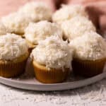 Coconut cupcakes sprinkled with coconut flakes on a round plate set on a white wood board.