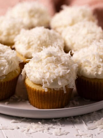 Coconut cupcakes sprinkled with coconut flakes on a round plate set on a white wood board.