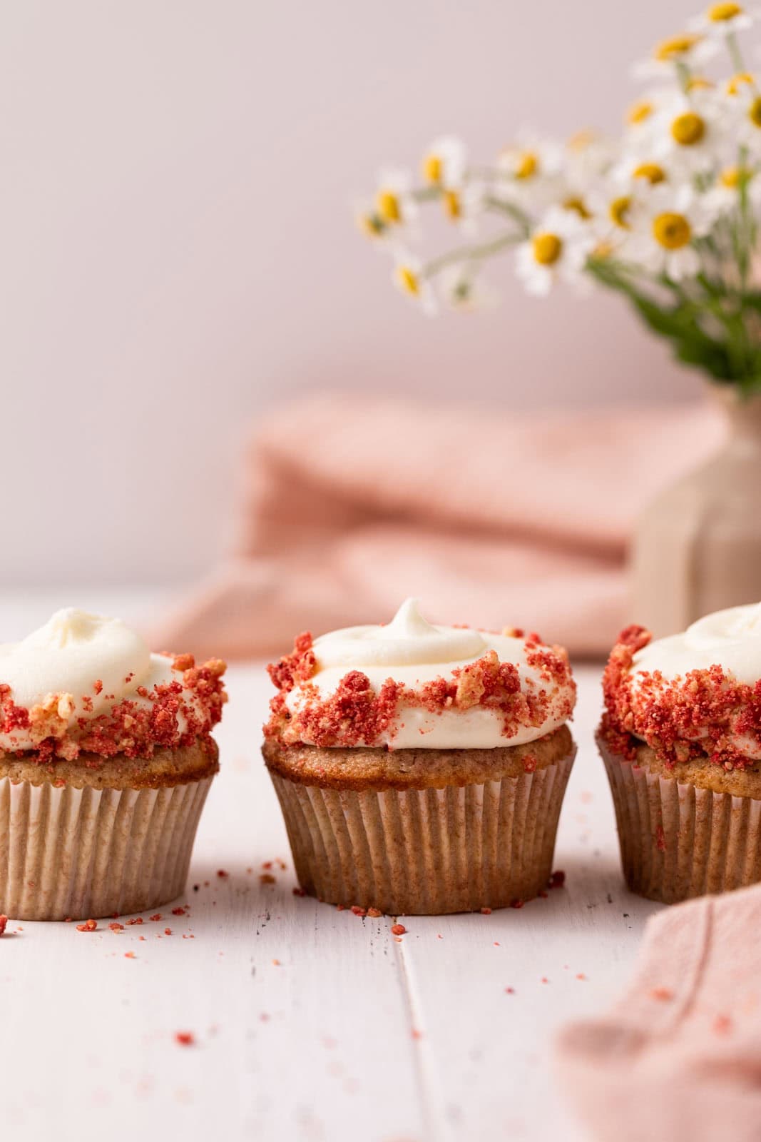 Three strawberry crunch cupcakes with cream cheese frosting lined up on a white wood board with flowers in the background.