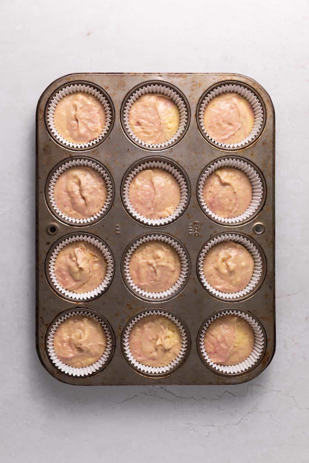 Cupcake batter divided into a 12 count muffin tin with white cupcake liners set on a flat surface.