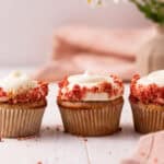 Three strawberry crunch cupcakes with cream cheese frosting lined up on a white wood board with flowers in the background.
