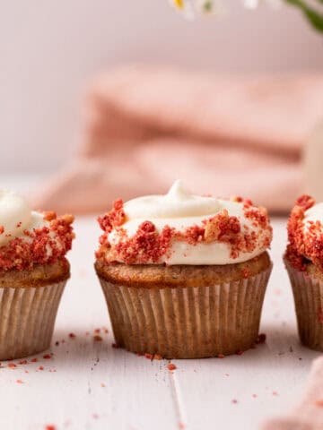 Three strawberry crunch cupcakes with cream cheese frosting lined up on a white wood board with flowers in the background.