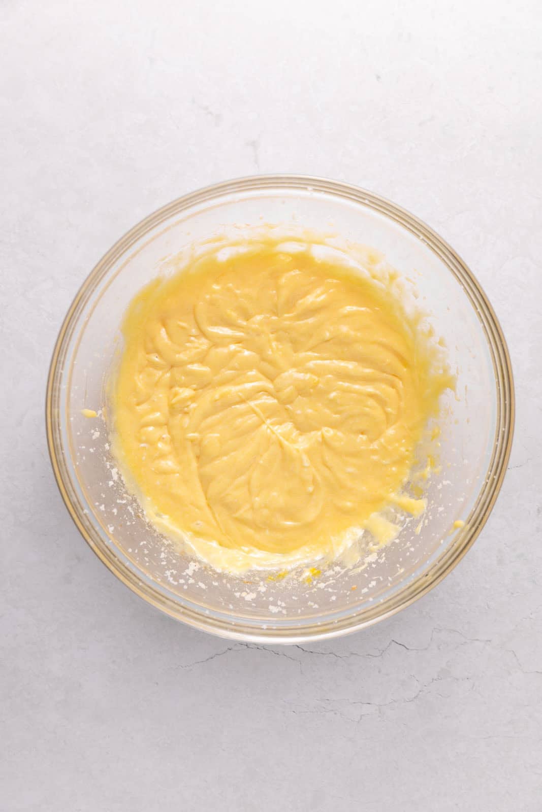 Sour cream mixed into creamed butter and sugar mixture with eggs and vanilla in a glass mixing bowl set on a flat surface.