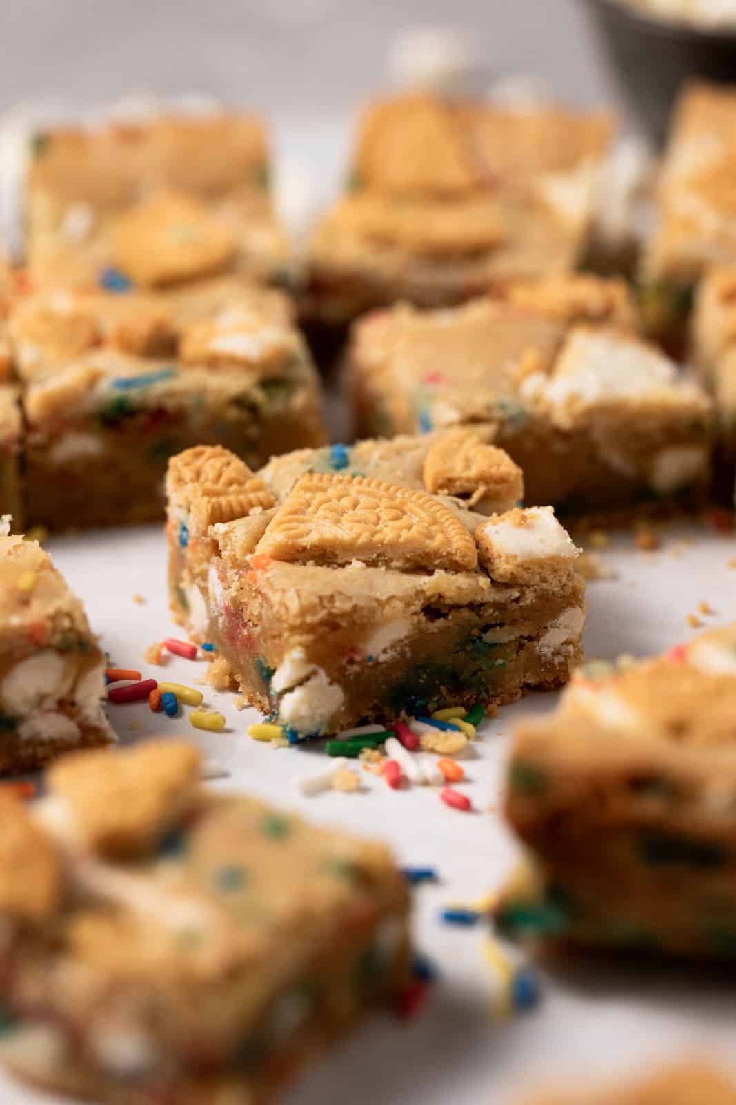 Close up of birthday cake blondie square set on a flat surface surrounded by more squares and sprinkles.
