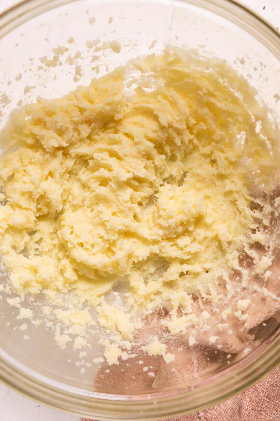 A glass bowl containing a creamy, pale yellow mixture of butter and sugar, partially mixed together, sitting on a light pink cloth.