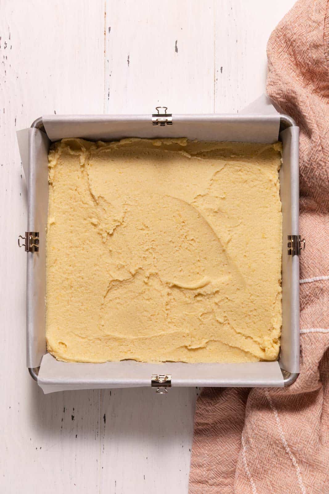 Lemon sugar cookie dough spread into a 8-inch square baking pan set on a white wooden surface.