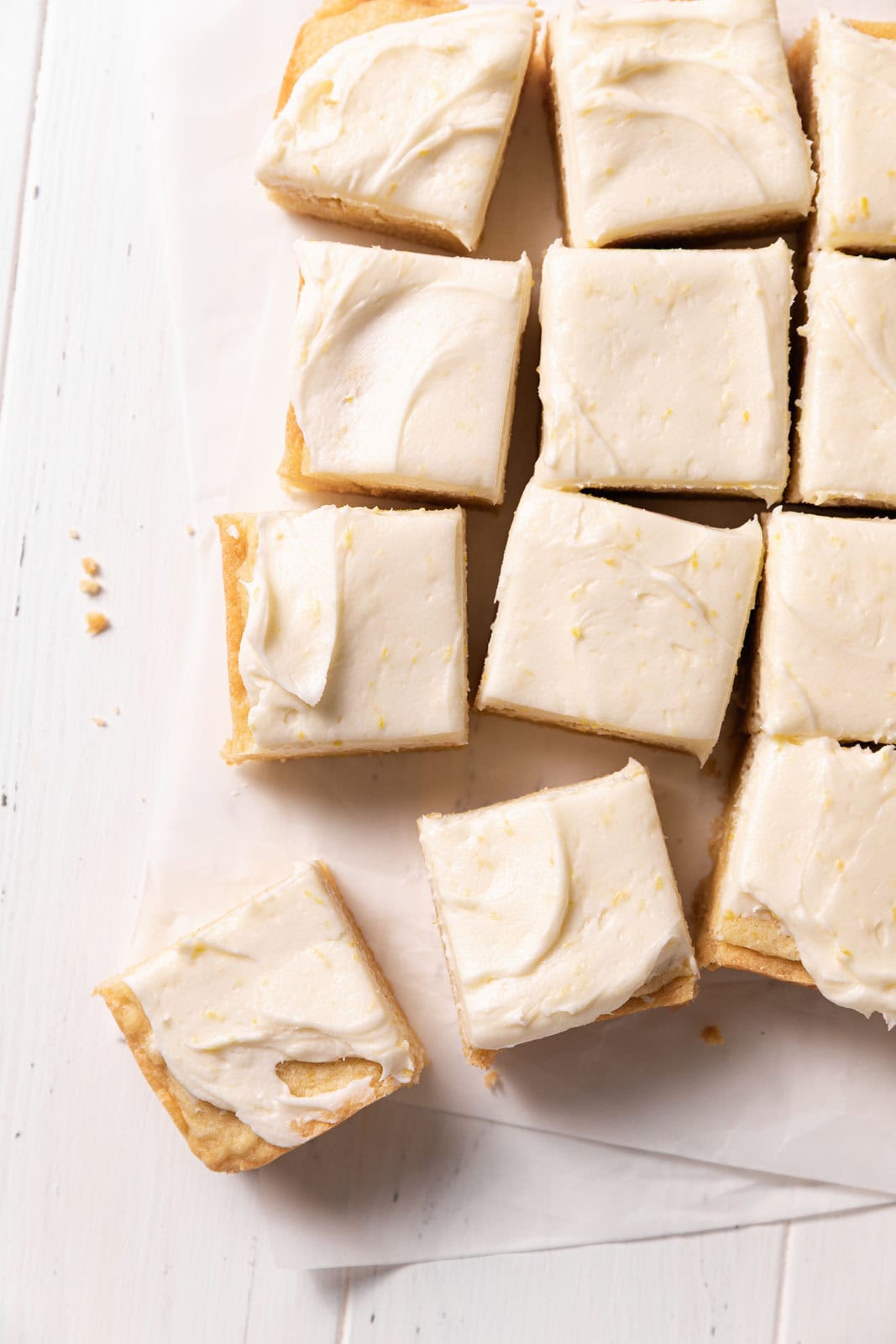 Frosted lemon sugar cookie bars cut into squares and arranged on parchment paper atop a white wooden surface.