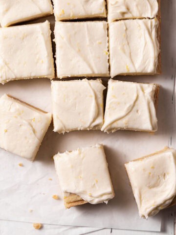 Frosted lemon sugar cookie bars cut into squares and arranged on parchment paper atop a white wooden surface.