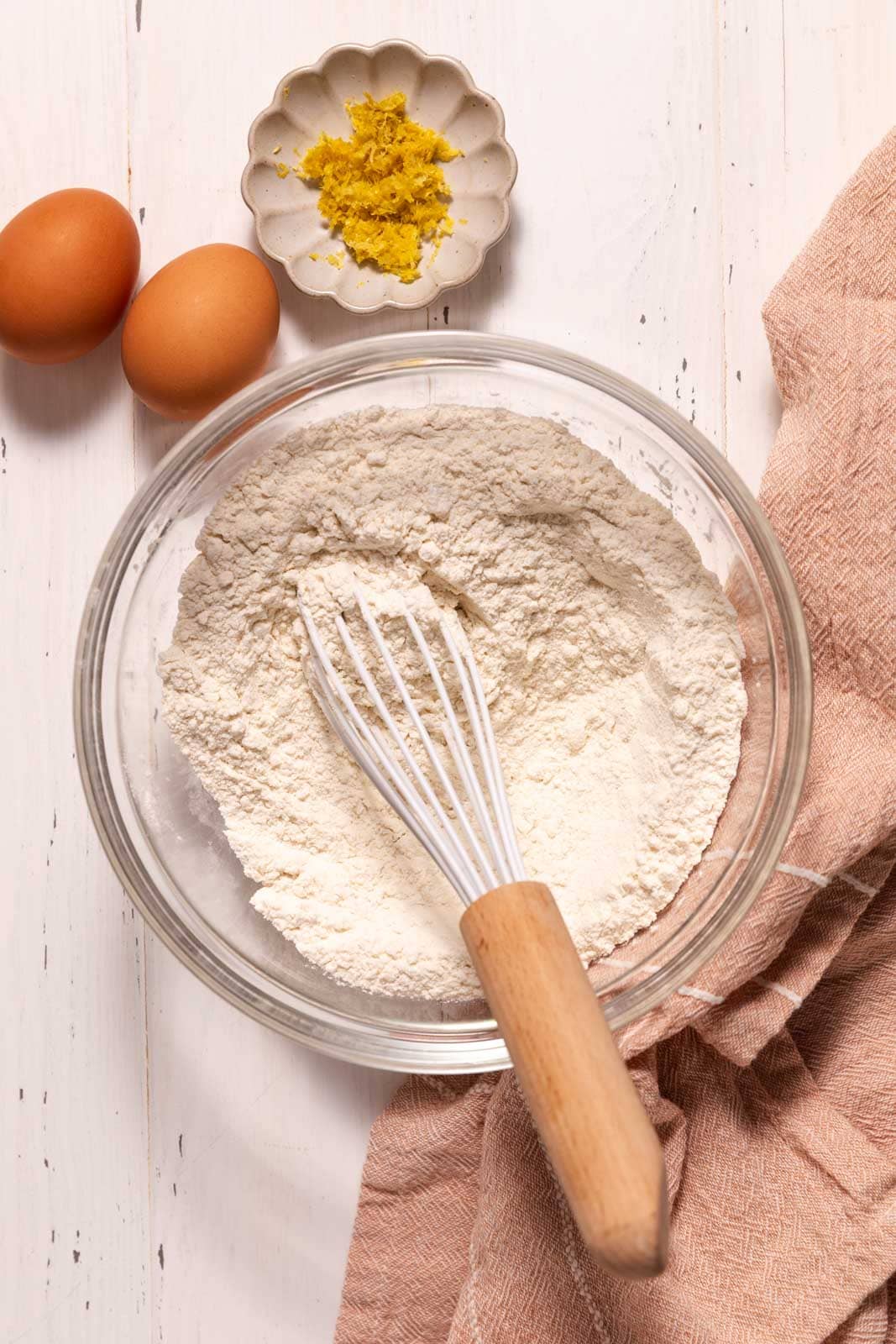 A glass bowl with flour and a whisk, two brown eggs, a small dish of lemon zest, and a pinkish kitchen towel on a white wooden surface.
