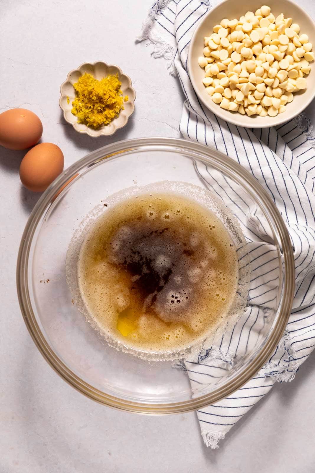 Overhead shot of brown butter in a glass mixing bowl set on a white board with a striped linen napkin, two eggs, lemon zest, and white chocolate to the sides.