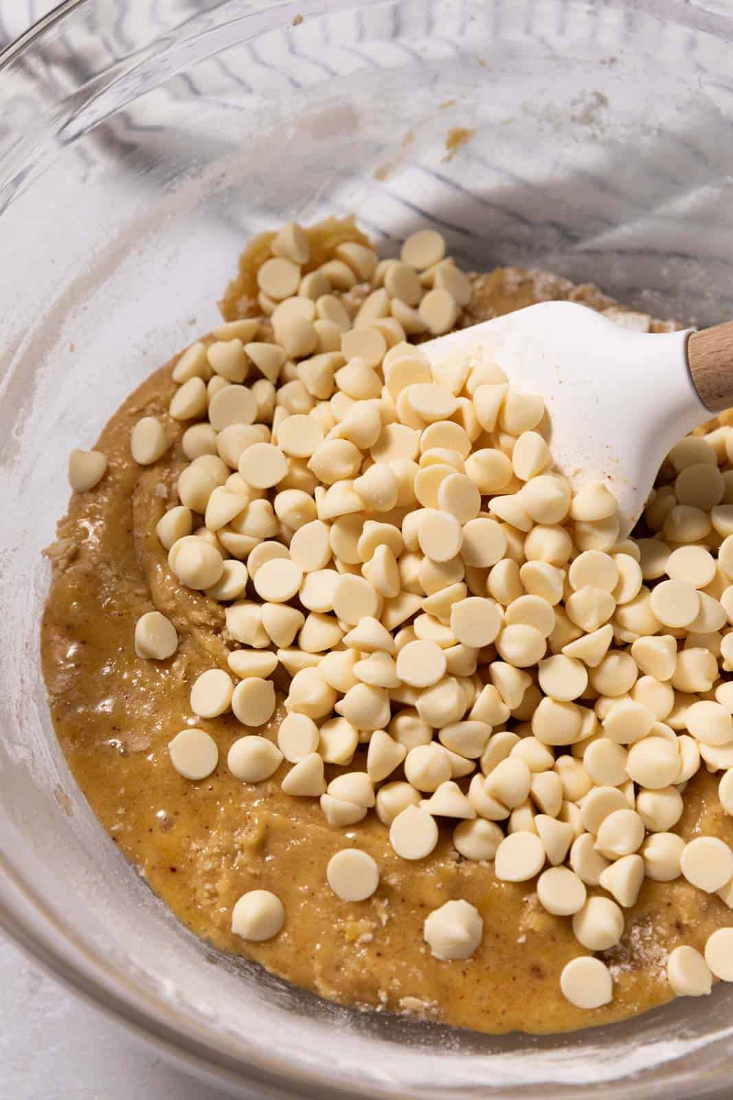 Close up of mixed cookie dough in a glass mixing bowl with a white rubber spatula and white chocolate chips over the dough.