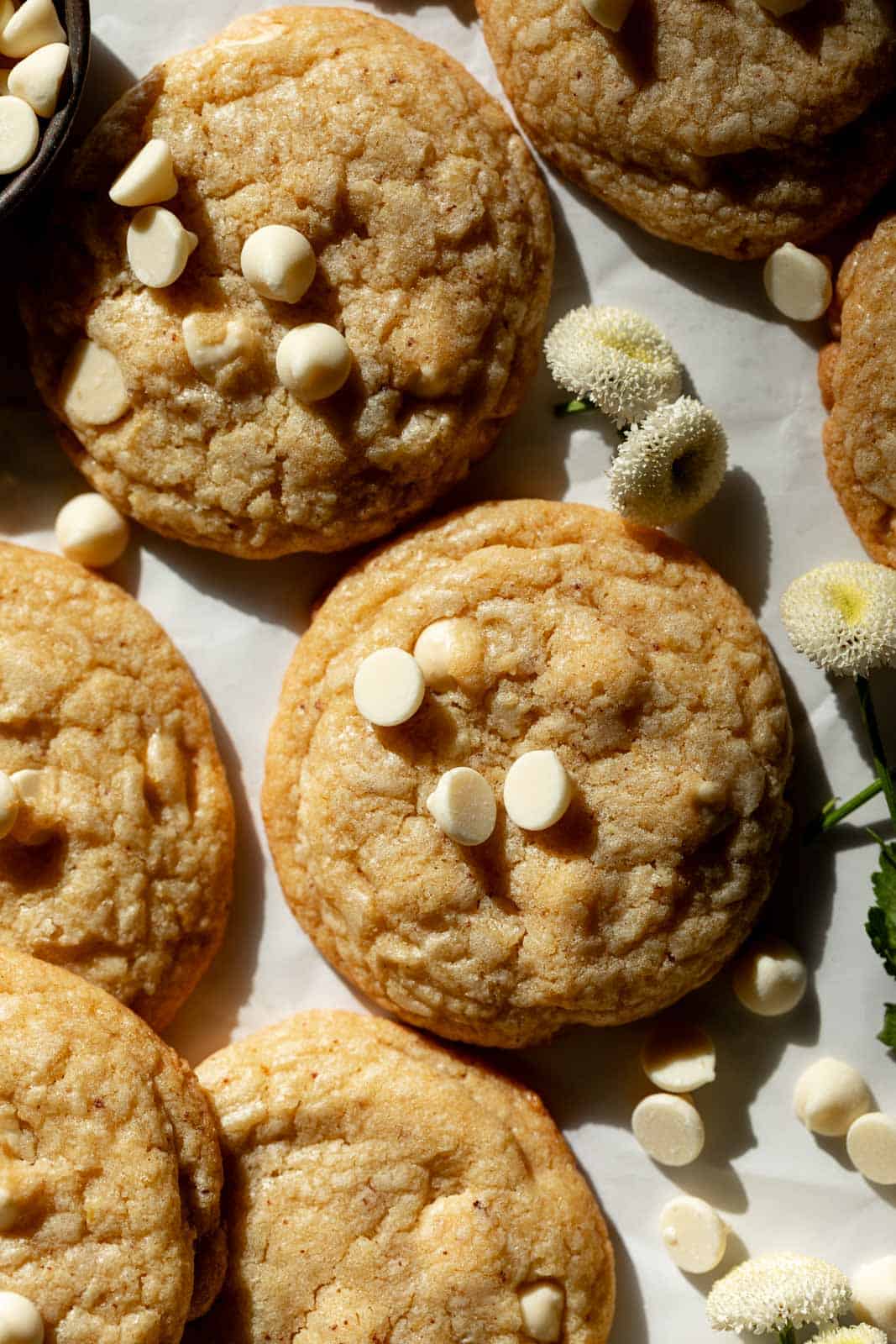 Overhead shot of lemon white chocolate chip cookies set on white parchment paper with small white flowers scattered around.