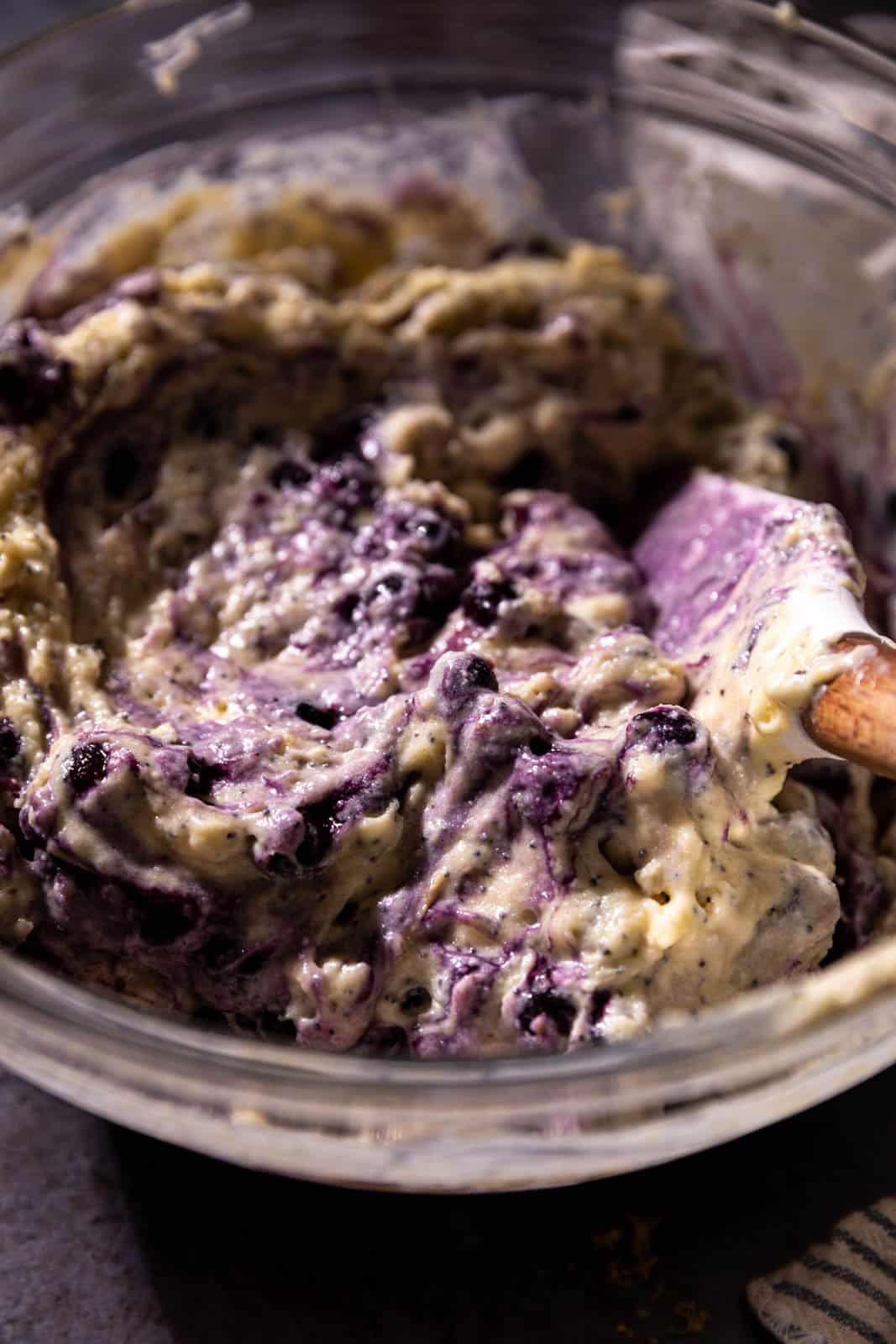 Wild blueberry and lemon muffin batter mixed with a white rubber spatula in a glass mixing bowl.