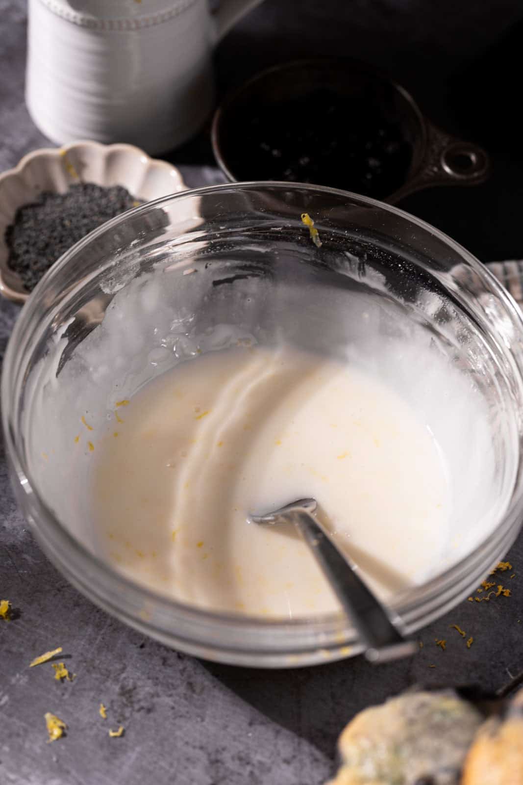 Whisk lemon glaze in a glass mixing bowl with a fork and a small dish of poppy seeds in the background.