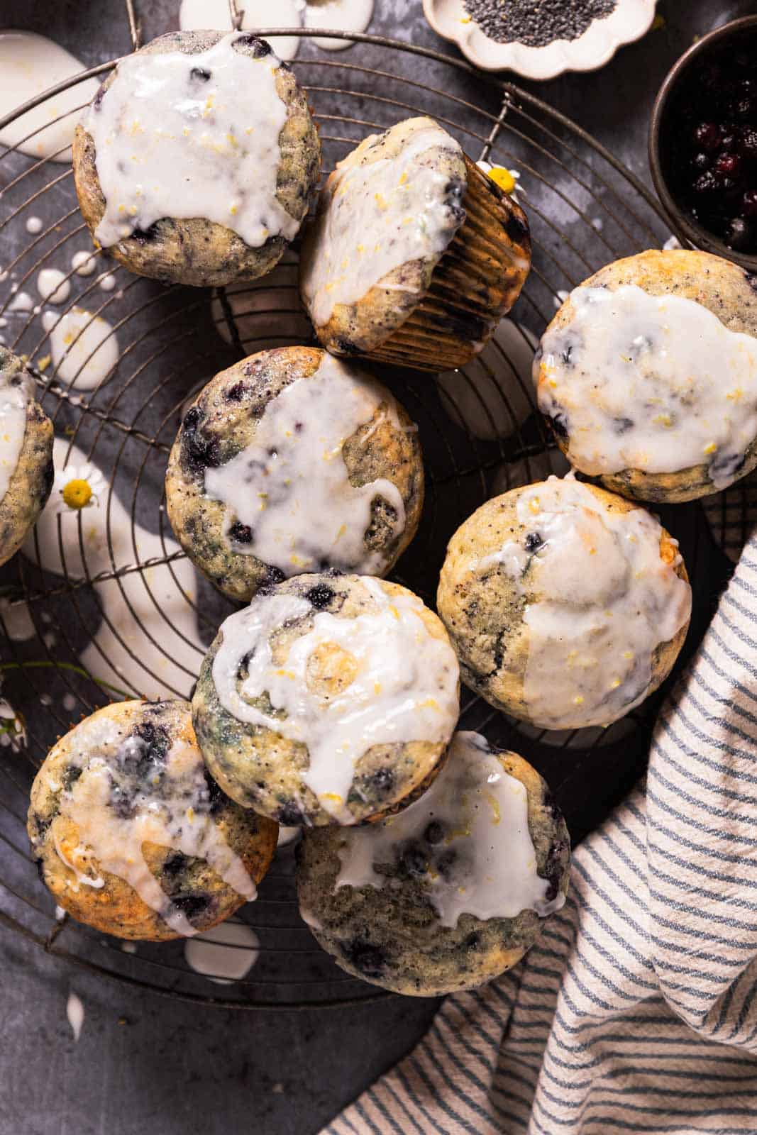 Overhead shot of a wild blueberry lemon poppy seeds muffins with a lemon glaze stacked on a round wire cooling rack with a striped linen on the side.