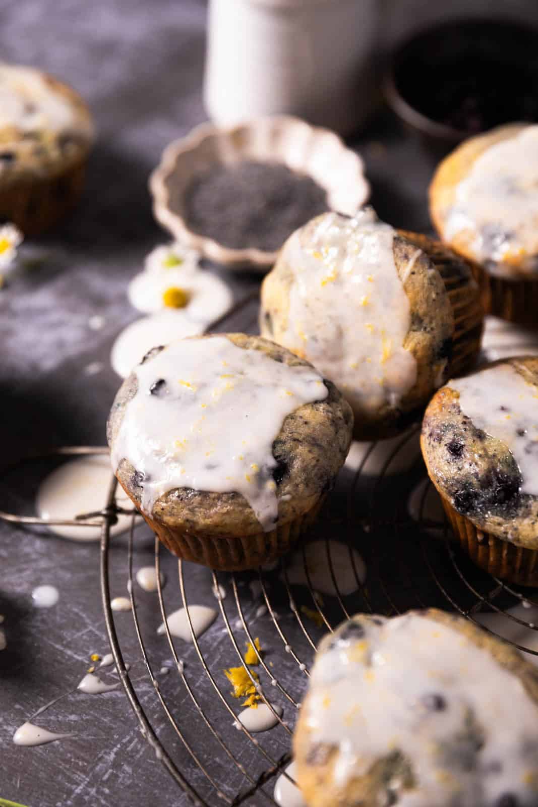 Wild blueberry lemon poppy seed muffins with a lemon glaze arranged randomly on a round wire rack set on a dark backdrop.