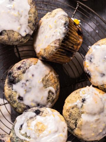 Overhead shot of a wild blueberry lemon poppy seeds muffins with a lemon glaze stacked on a round wire cooling rack with a striped linen on the side.