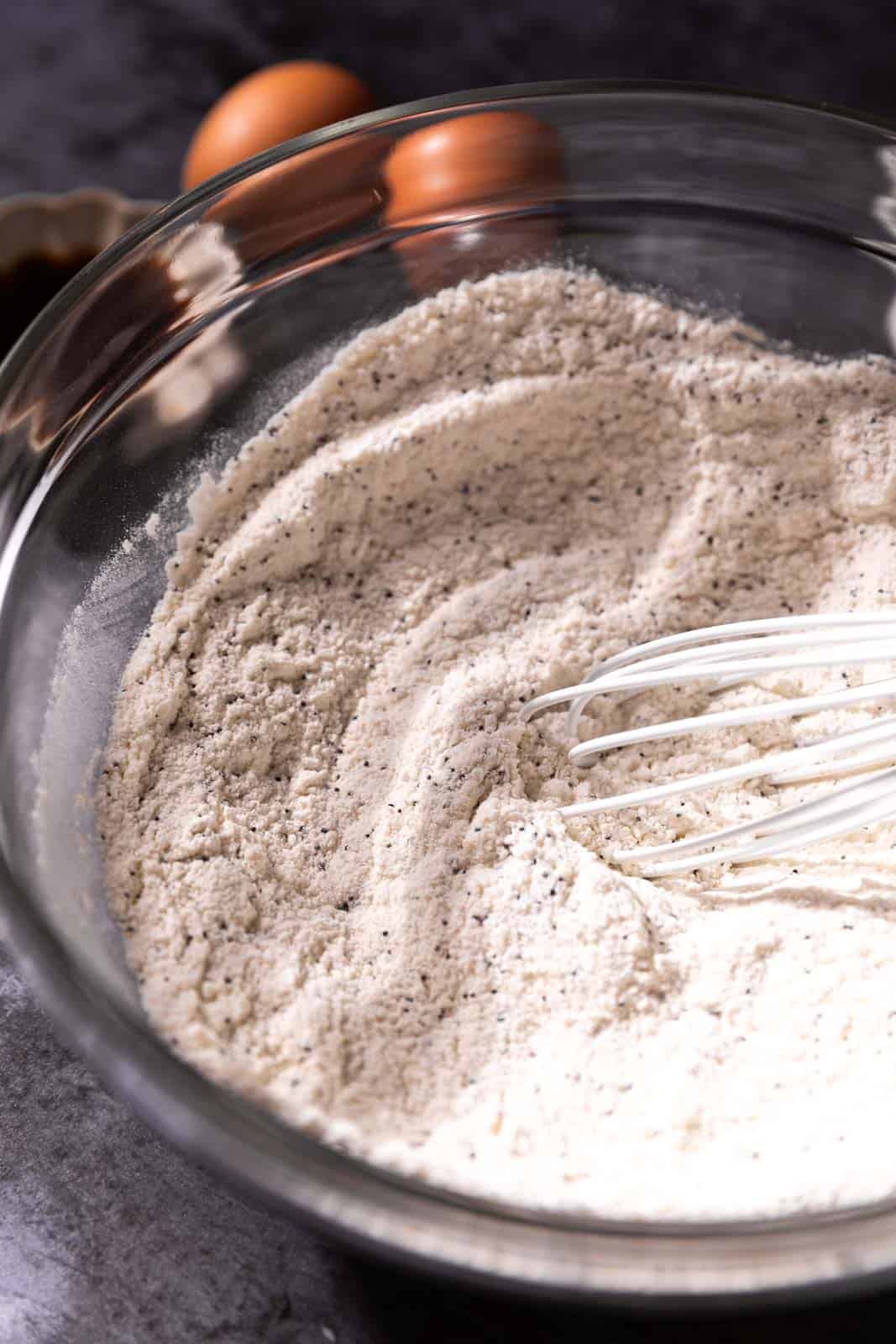 Whisked dry ingredients in a glass bowl with a white whisk and two eggs in the background.