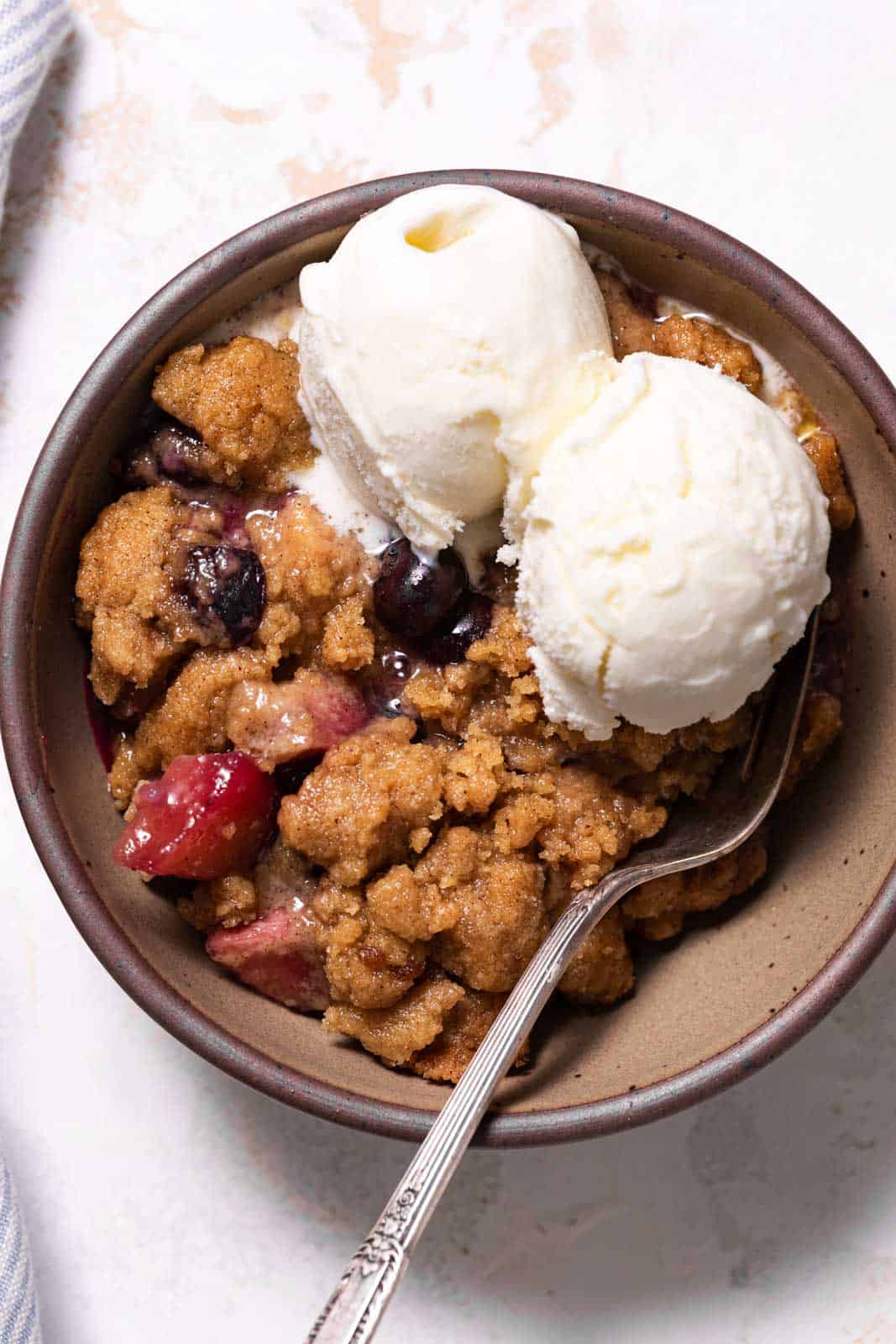 Overhead shot of a bowl of apple blueberry crumble with two scoops of vanilla ice cream and a fork.