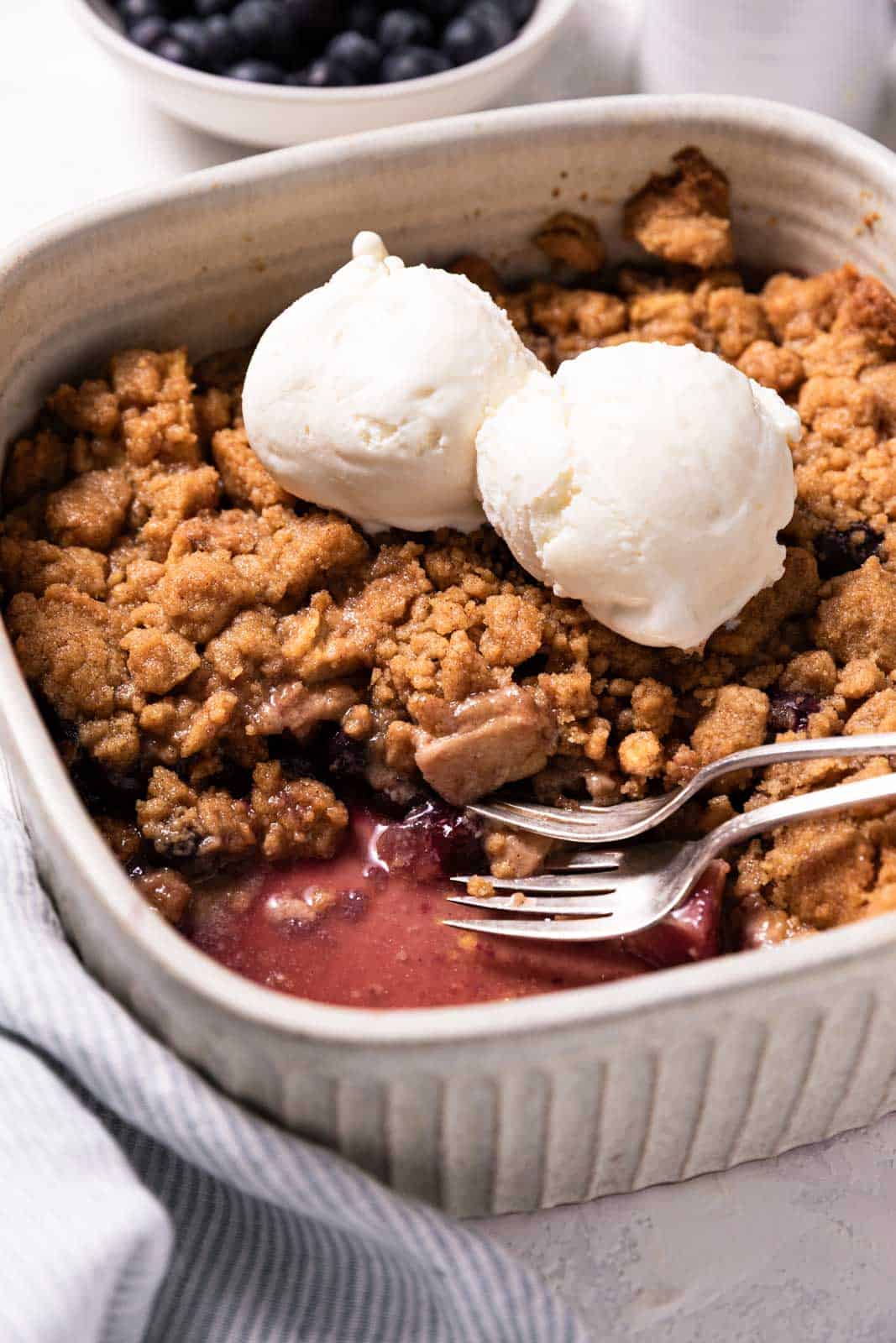 A deep square baking dish with apple blueberry crumble topped with two scoops of vanilla ice cream and two forks.