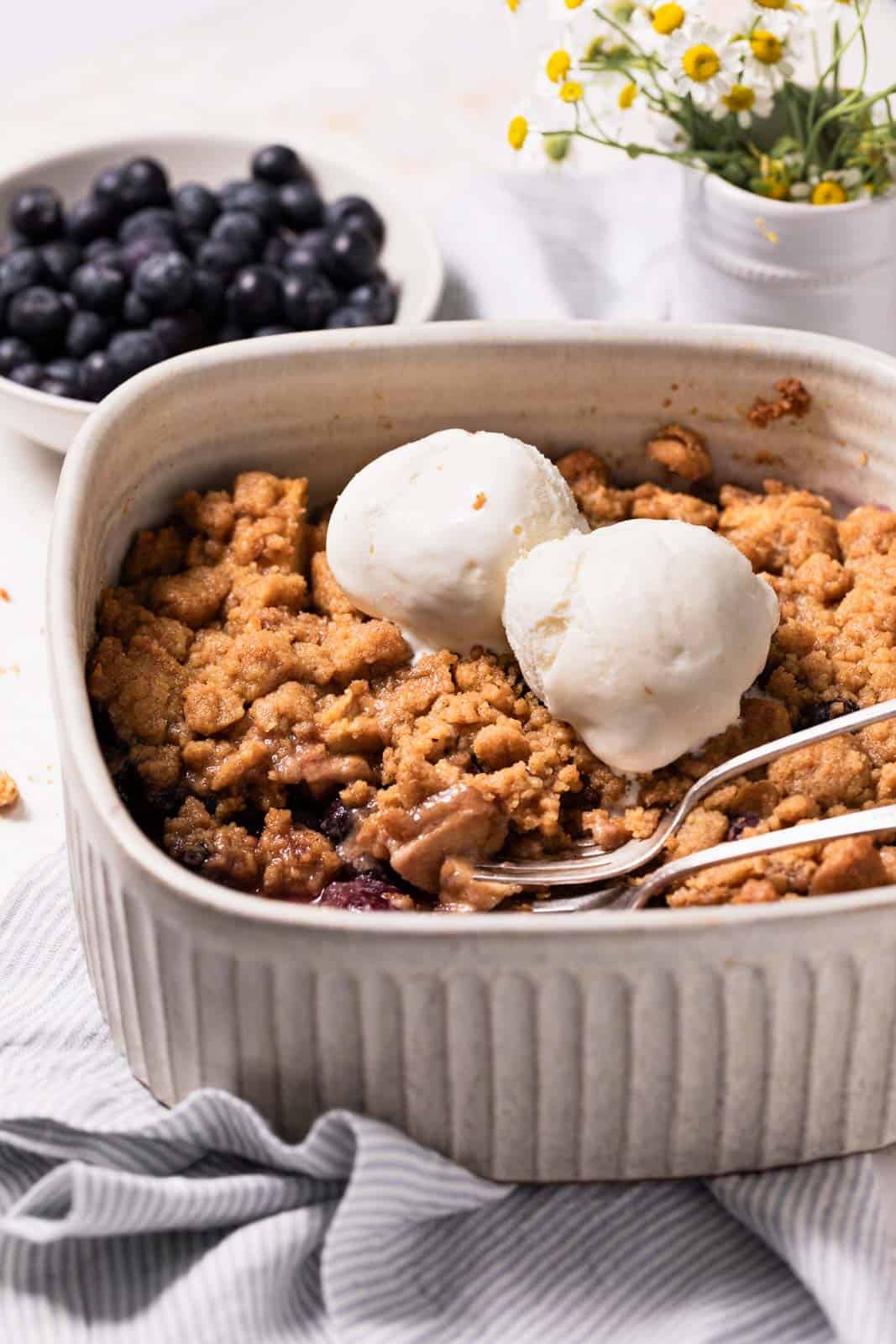 A deep square baking dish with apple blueberry crumble topped with two scoops of vanilla ice cream and two forks and a bowl of blueberries in the background.