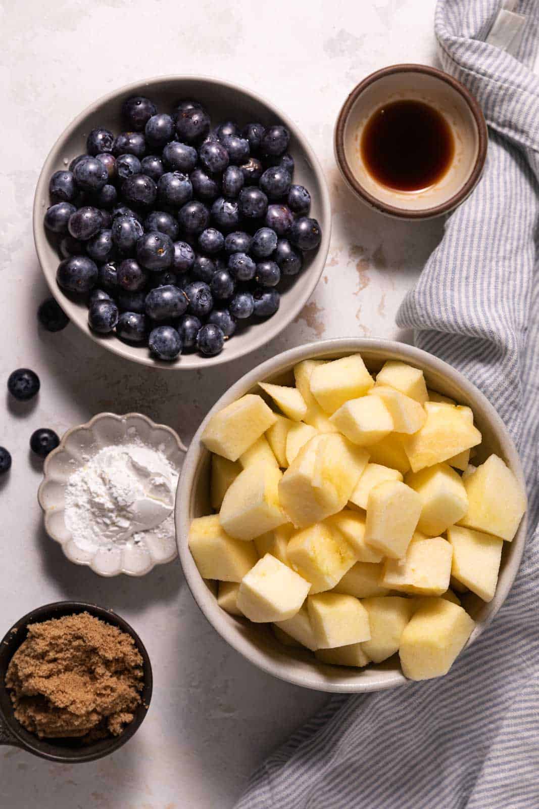 Filling ingredients for apple blueberry crumble in bowls set on a flat wood surface with a striped linen.
