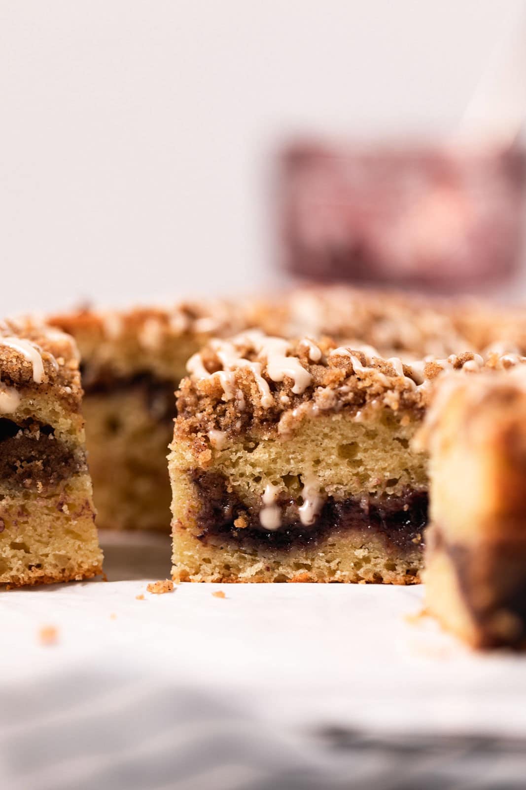 Close up of square cut blackberry coffee cake drizzled with vanilla glaze set on white parchment paper.