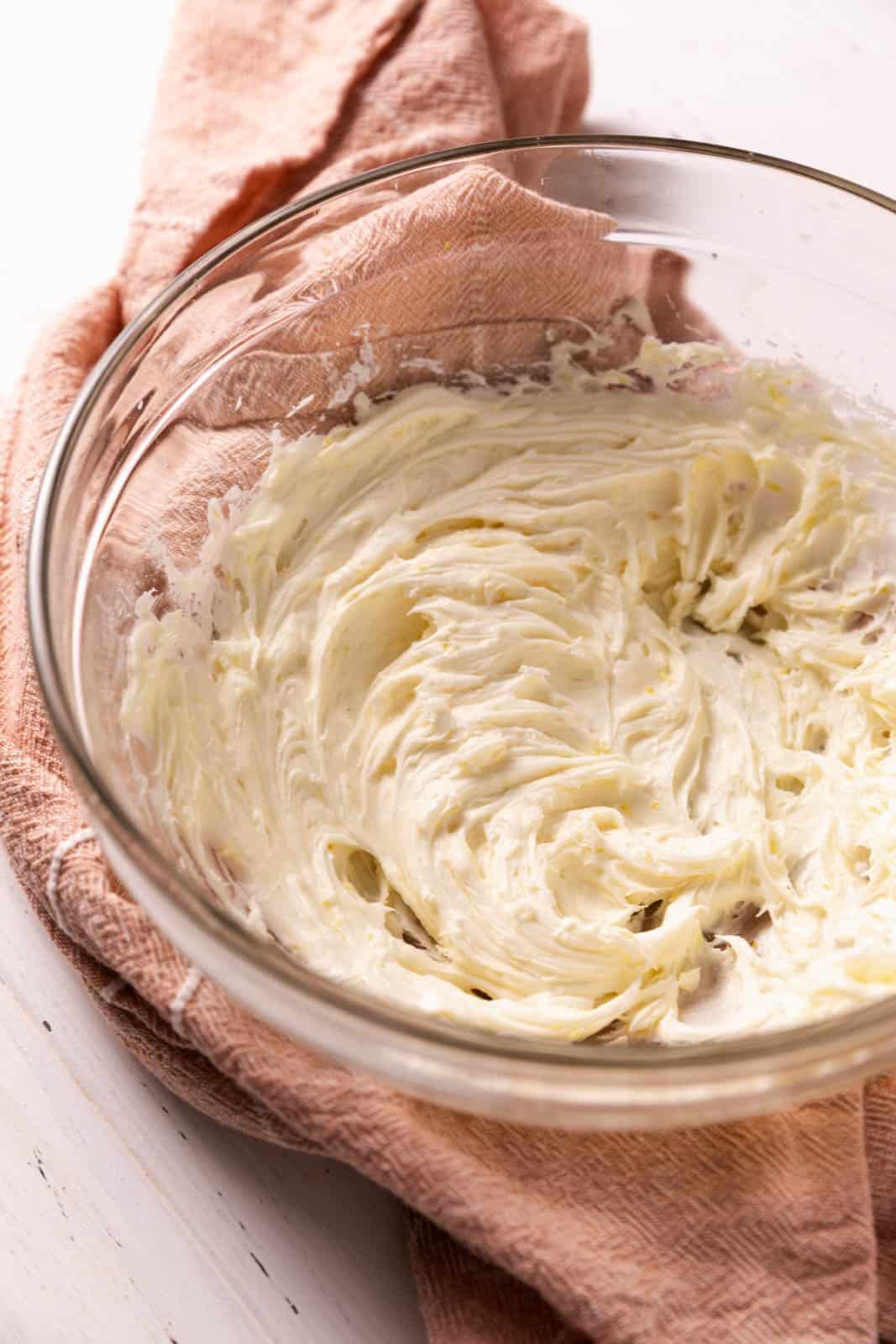 Lemon cream cheese frosting in a glass mixing bowl set on a pink towel.