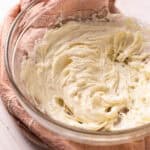 Lemon cream cheese frosting in a glass mixing bowl set on a pink towel.