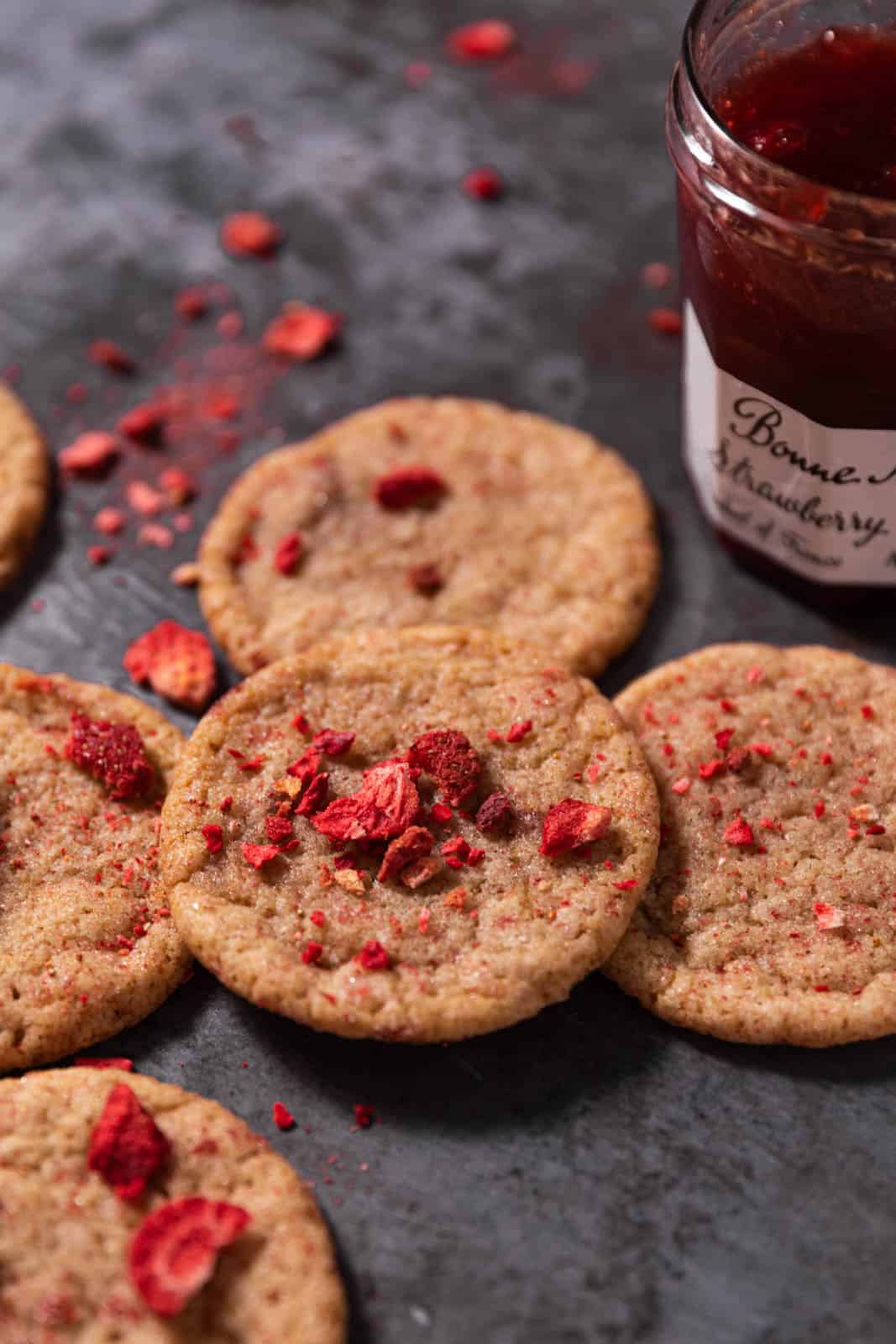 Strawberry sugar cookies topped with freeze dried strawberries arranged on a dark backdrop with a jar of strawberry jam.