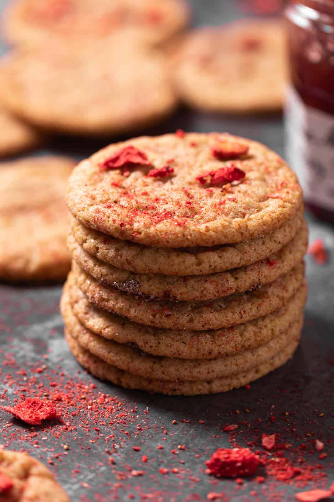 Stack of strawberry sugar cookies with freeze-dried strawberries on a dark backdrop with a jar of strawberry jam.
