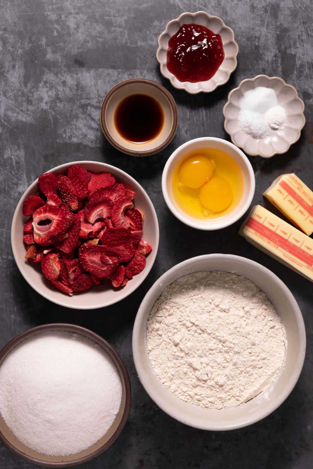 Strawberry sugar cookies ingredients in bowls on a flat dark backdrop.
