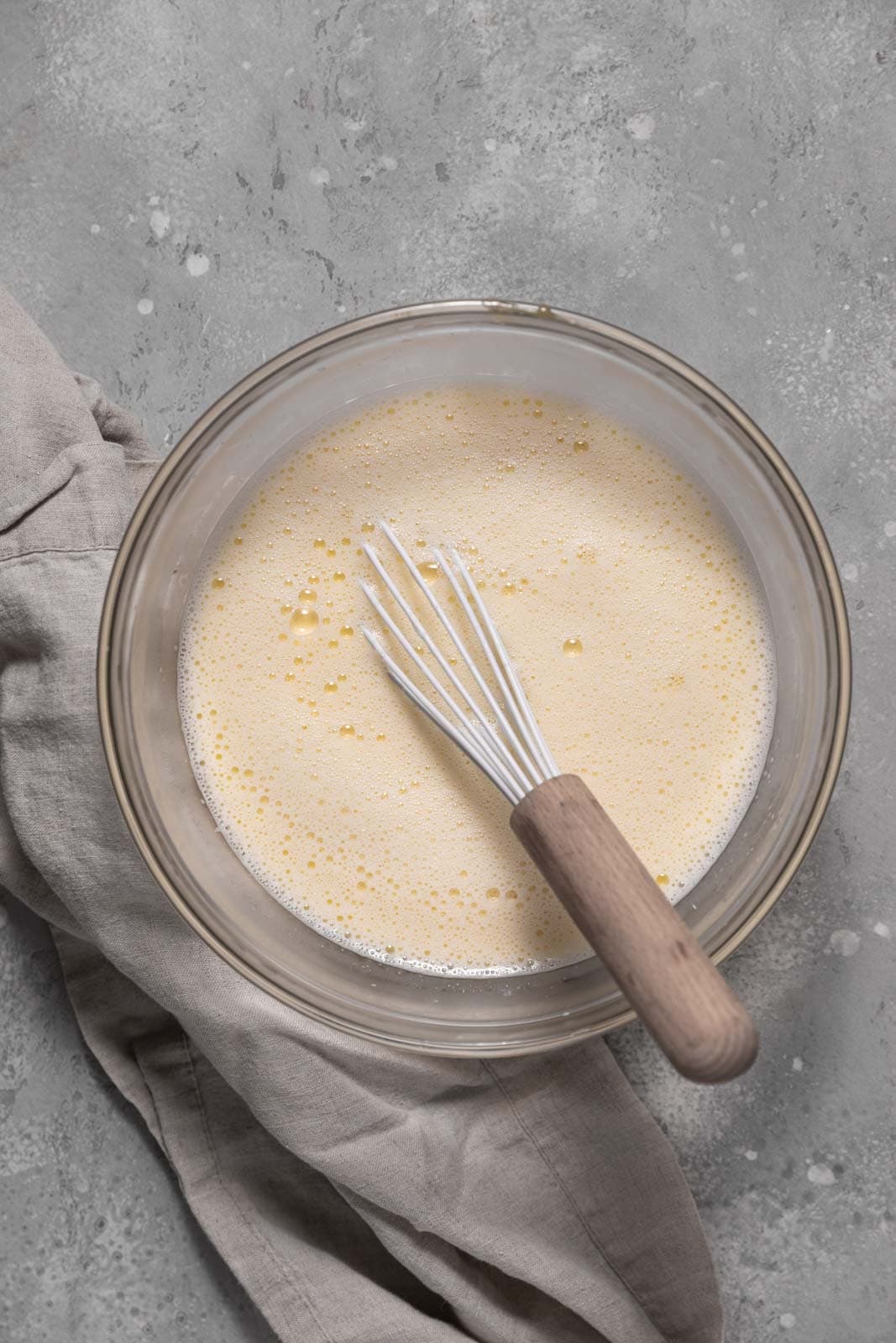 Whisked milk and cream with eggs and sugar in a glass mixing bowl set on a flat surface with a whisk.