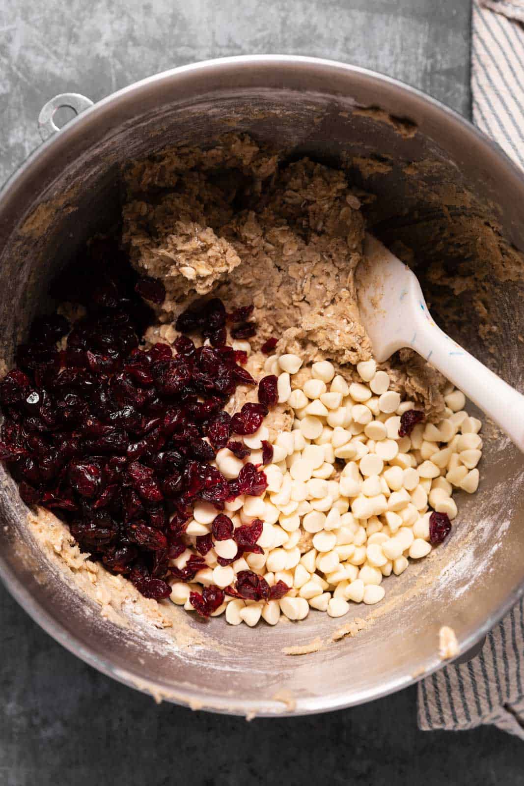 Oatmeal cookie dough in a metal stand mixer bowl with a white spatula with dried cranberries and white chocolate to be mixed in.