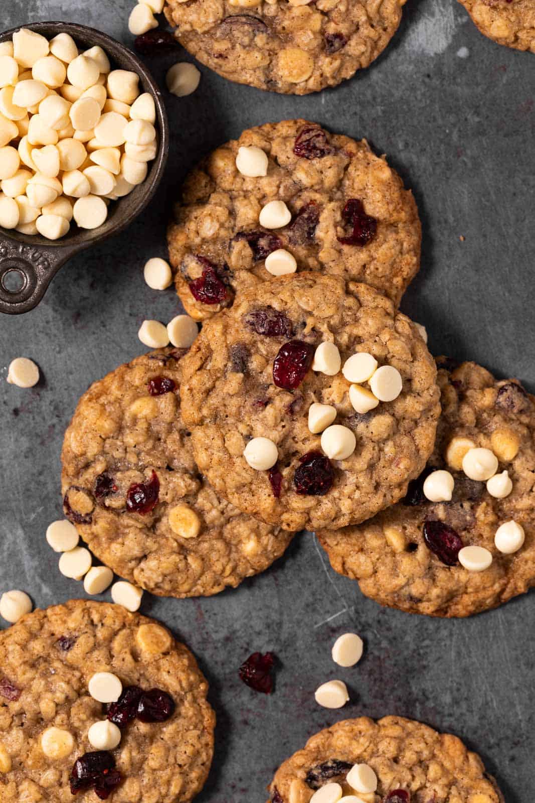 Overhead shot of white chocolate cranberry oatmeal cookies arranged on a flat surface with a cup of white chocolate chips.