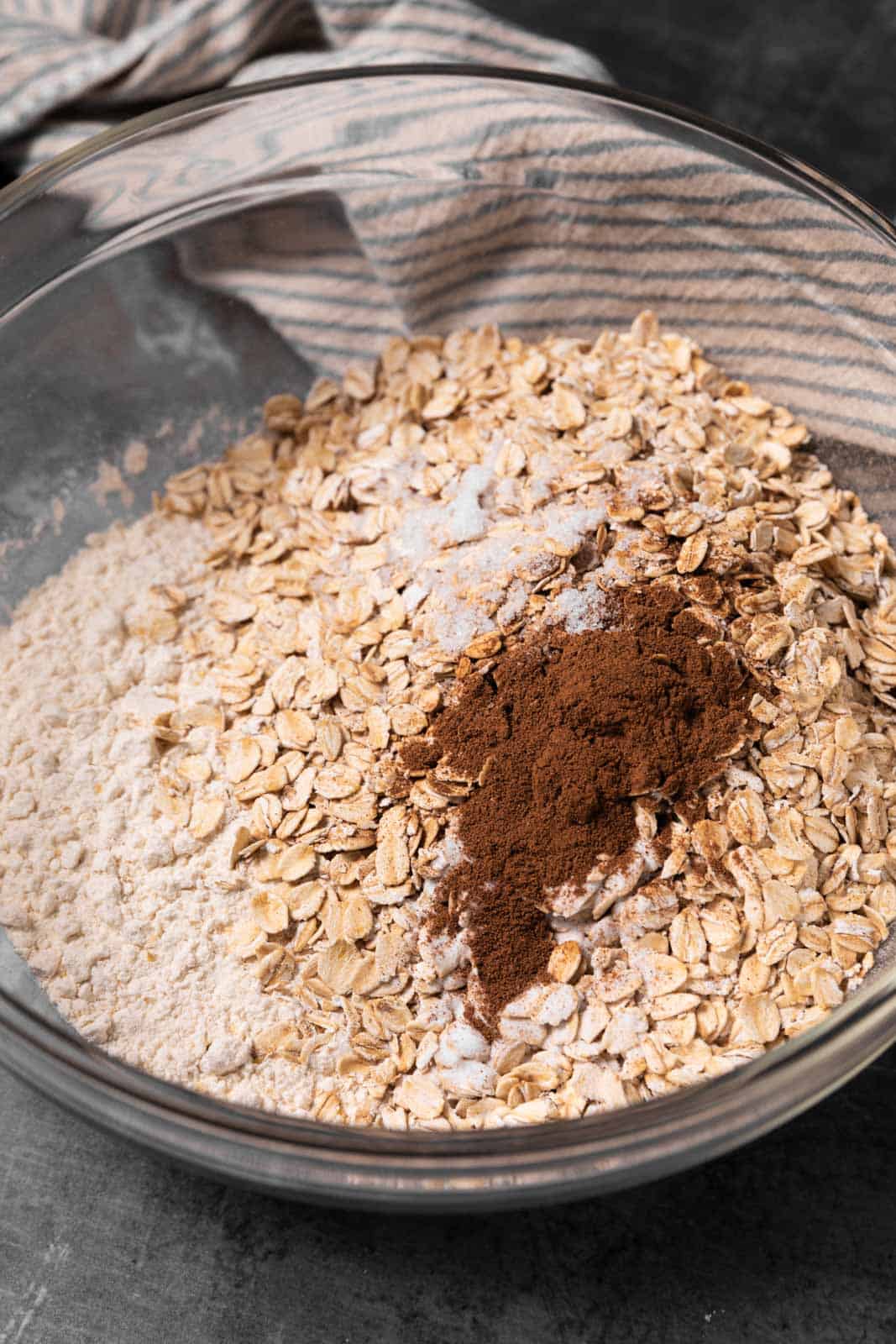 Dry ingredients for oatmeal cookies in a glass mixing bowl set on a flat surface with a linen napkin.