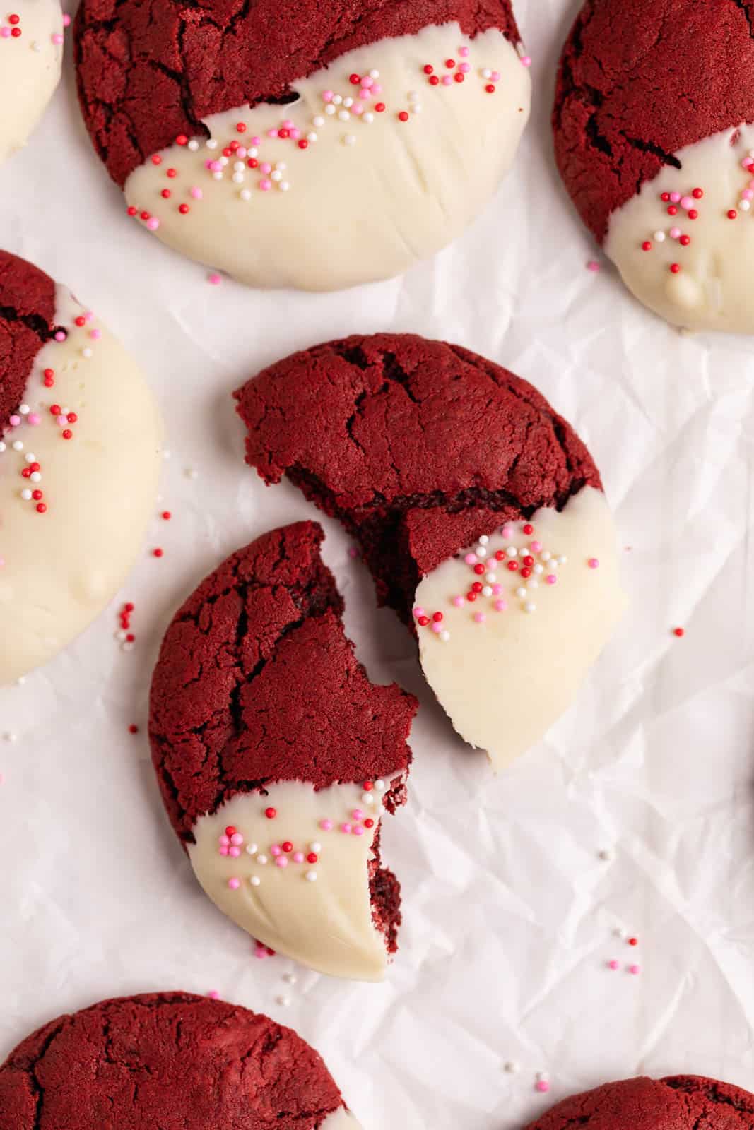Close up of red velvet cookie dipped in white chocolate with valentine colored perils and broken in half set on crinkled parchment paper.