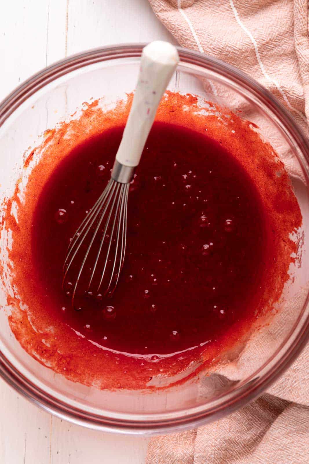 Whisked butter, sugar, egg, vanilla and red food coloring in glass mixing bowl set on a white wood board with pink linen napkin.