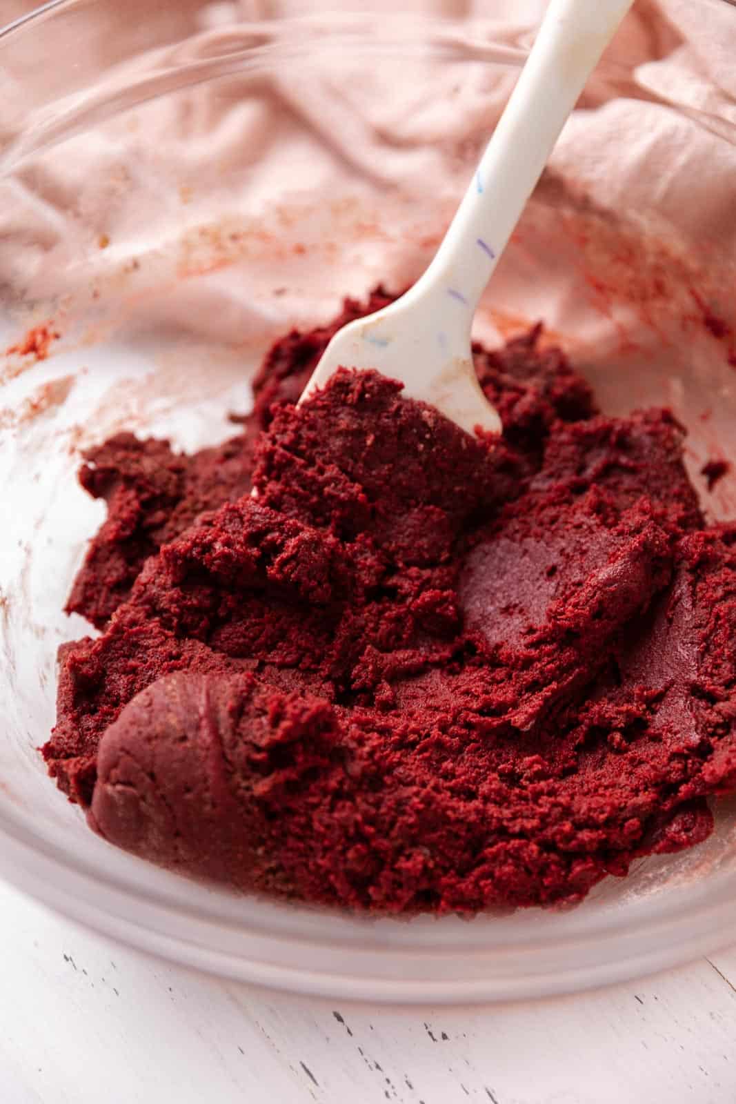 Red velvet cookie dough mixed in a glass mixing bowl with a rubber spatula set on a white wood board with pink linen.