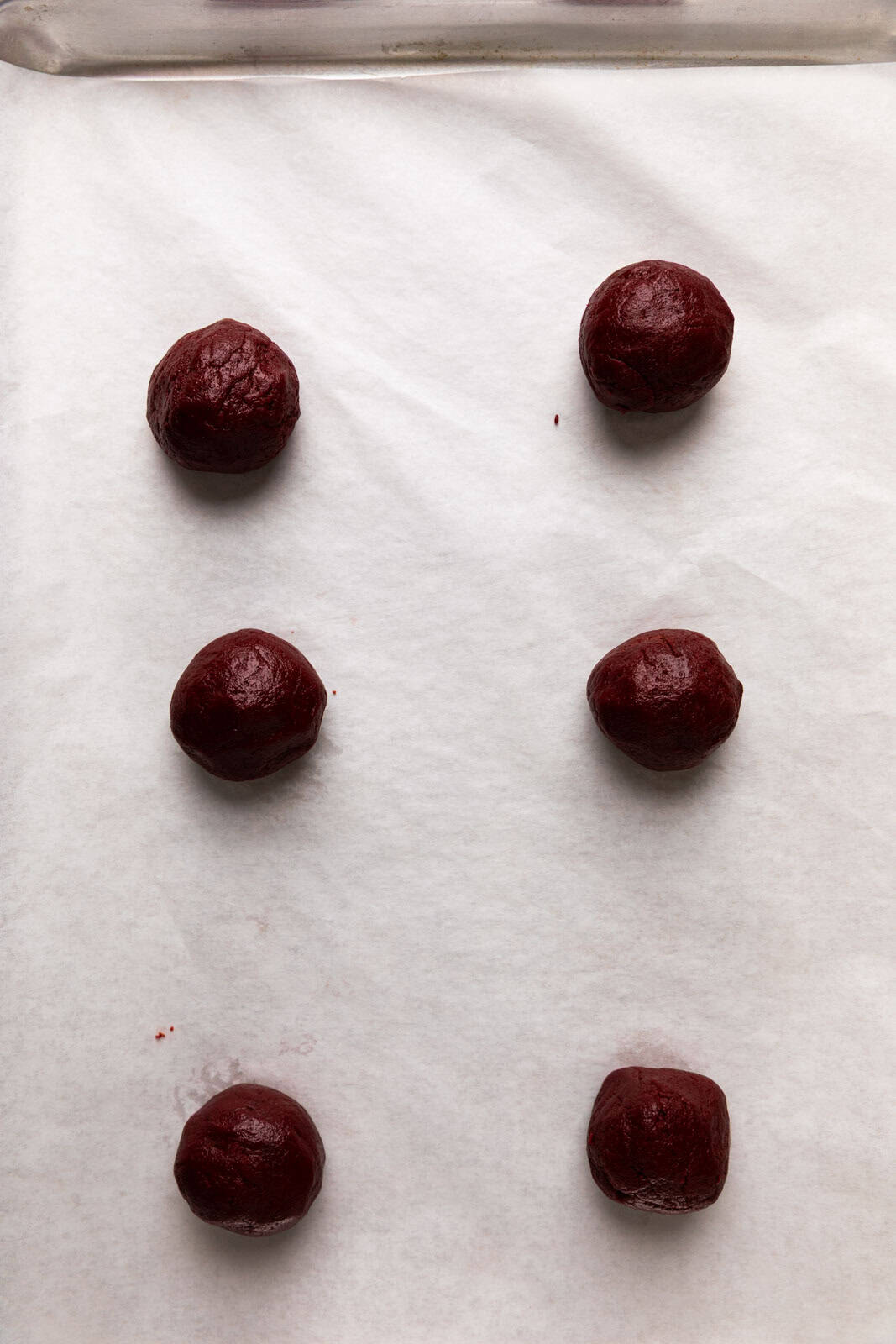 Six red velvet cookie dough balls spaced out on a parchment lined sheet pan.