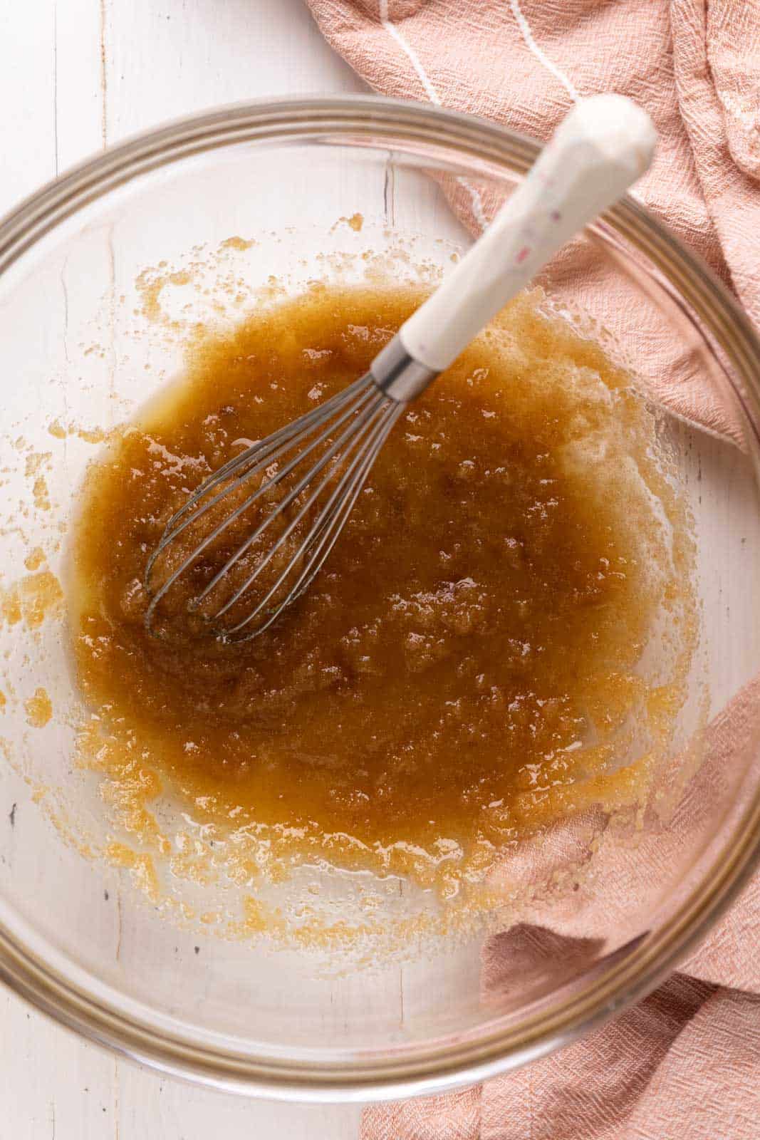 Whisked melted butter and sugar in a glass mixing bowl set on a white wood board with pick linen.