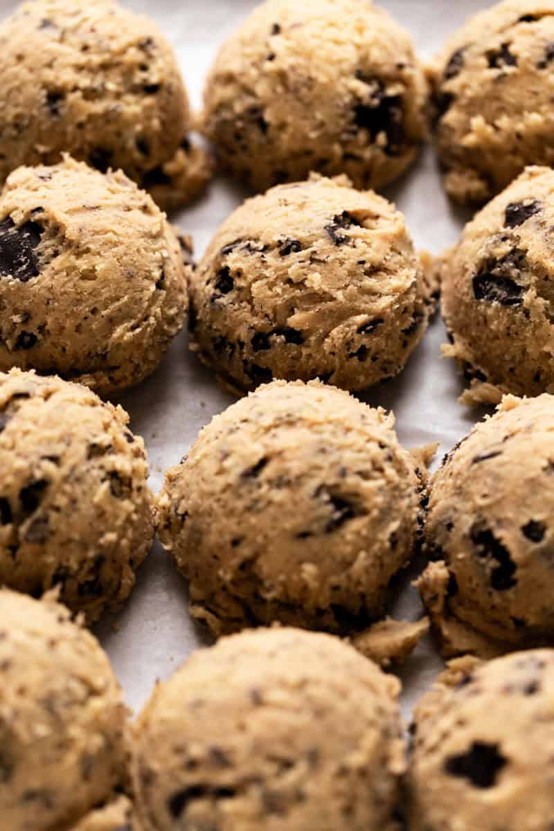 Chocolate chip cookies dough scooped and arranged side by side on a parchment lined sheet pan.
