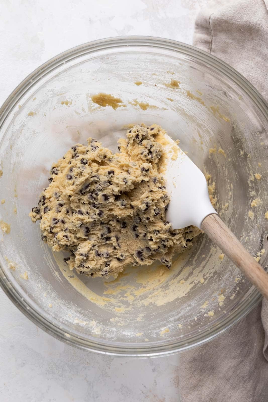 Edible chocolate chip cookie dough mixed in a glass mixing bowl with a white and wooden spatula.