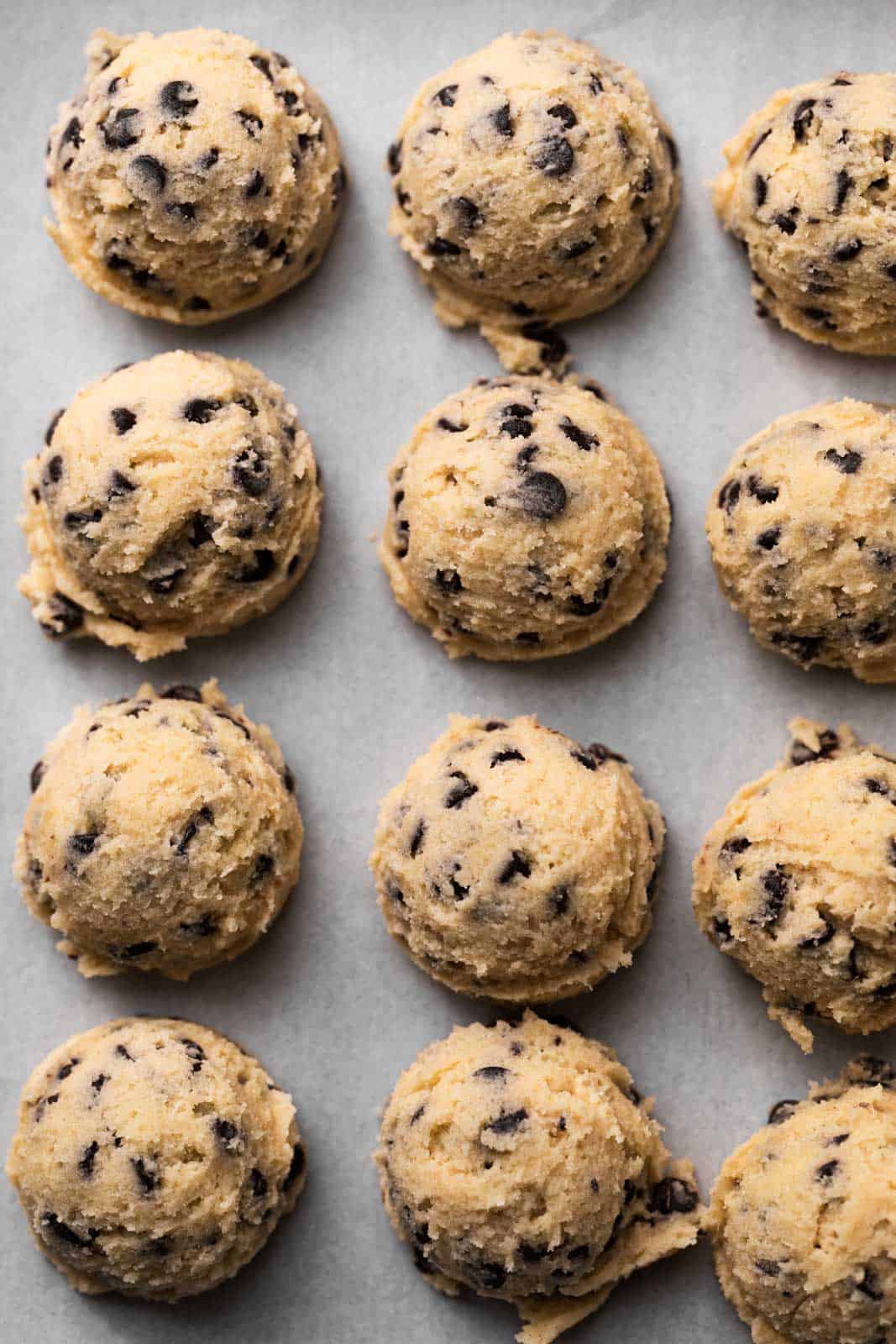Edible chocolate chip cookie dough scooped and spaced out in rows on a parchment-lined sheet pan.