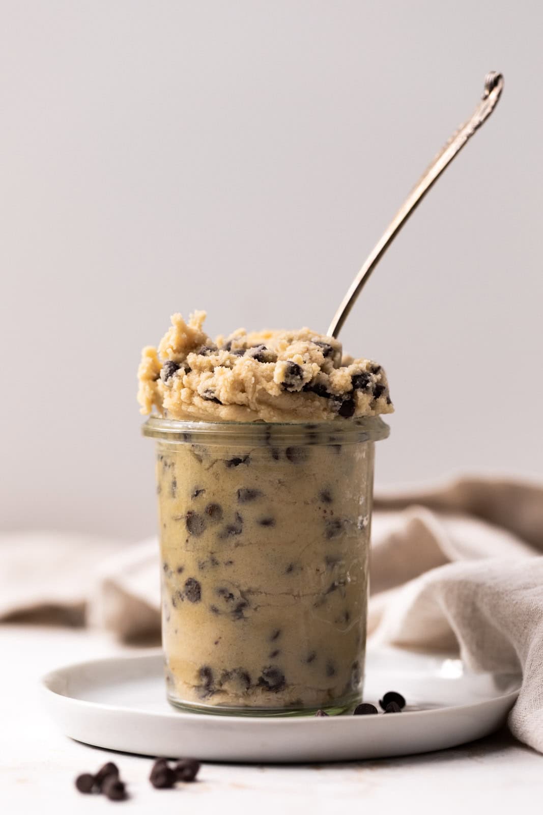 Edible chocolate chip cookie dough in a glass jar on a white plate with a spoon coming out of the cookie dough.